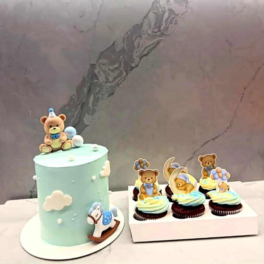 Pale blue teddy bear cake with clouds and a rocking horse, beside a box of teddy cupcakes